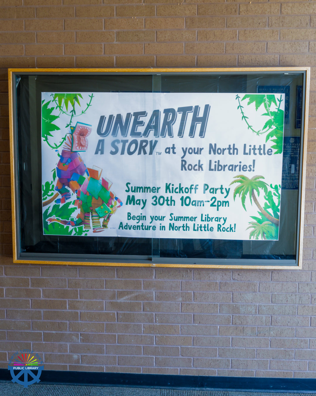 A sign behind glass reading Unearth a Story at your North Little Rock Libraries! Summer Kickoff Party May 30th 10am-2pm. Begin your Summer Library Adventure in North Little Rock.