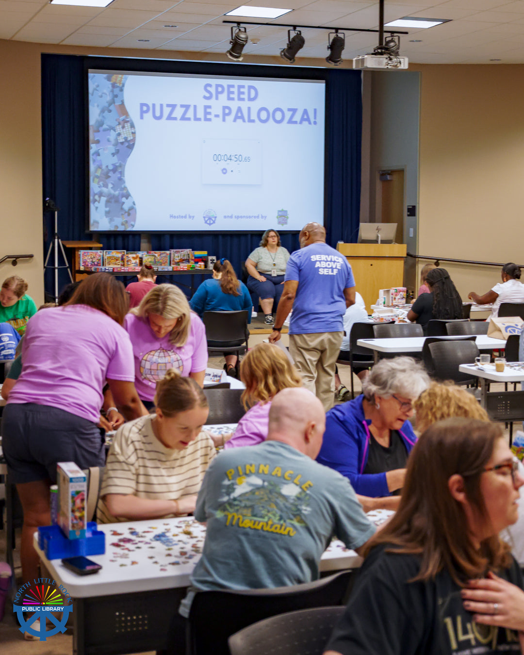Many people gathered around white tables work on colorful puzzles. A screen at the front of the room reads Speed Puzzle-Palooza! with a timer underneath.