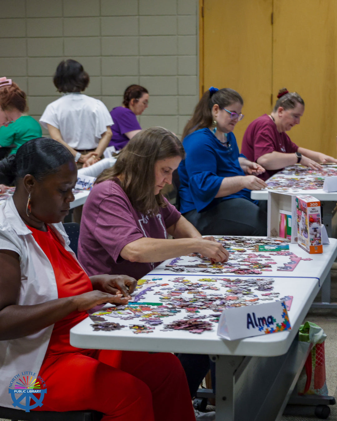 Individuals sit at white tables working on a jigsaw puzzle.