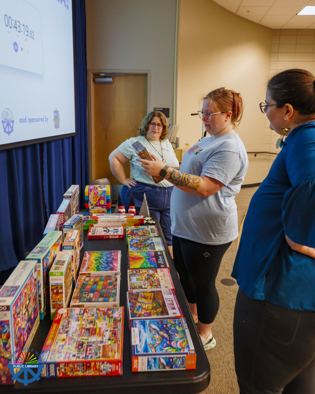A library staff member watches as two puzzle competitors select prizes from a table covered in colorful puzzles and puzzling accessories.
