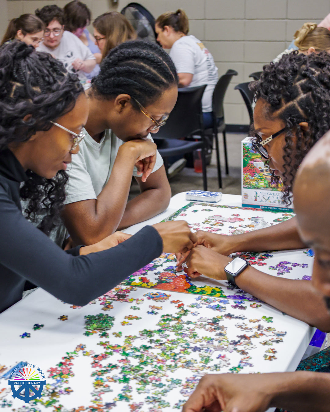 A group of 4 people lean over a table, working on a puzzle together. Other groups work similarly in the background.