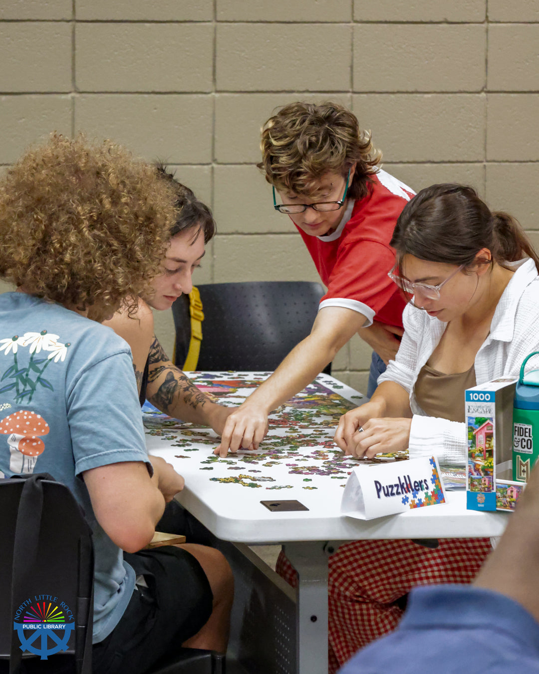A standing person points to a puzzle piece with 3 other seated people look at the pieces.