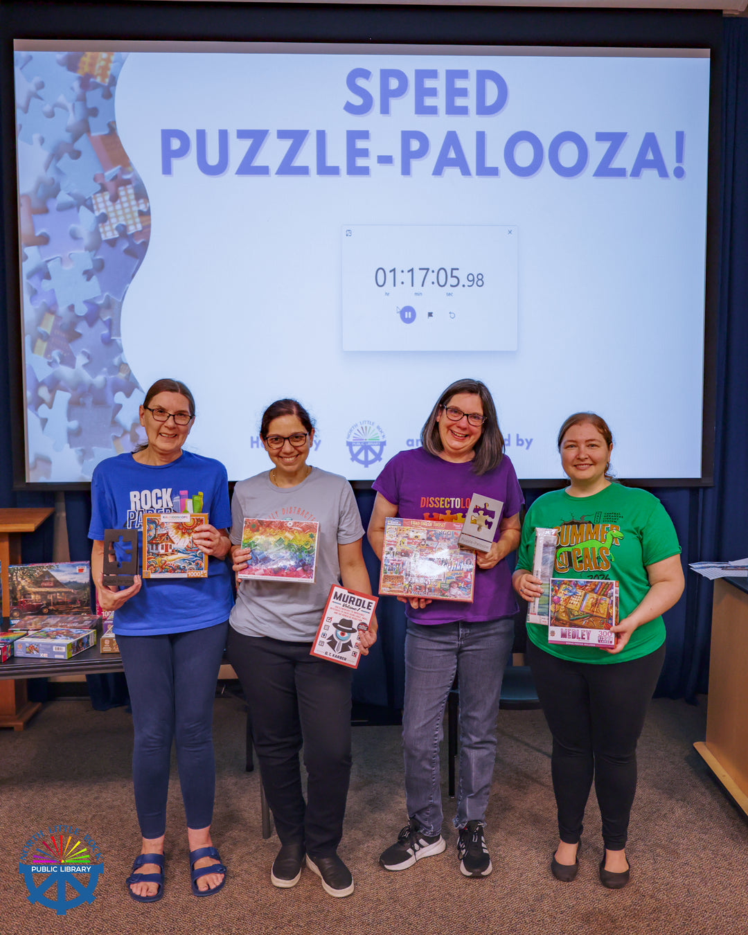 Four people holding puzzles and puzzle accessories pose in front of a screen reading Speed-Puzzle-Palooza! with a timer reading 01:17:05.98.