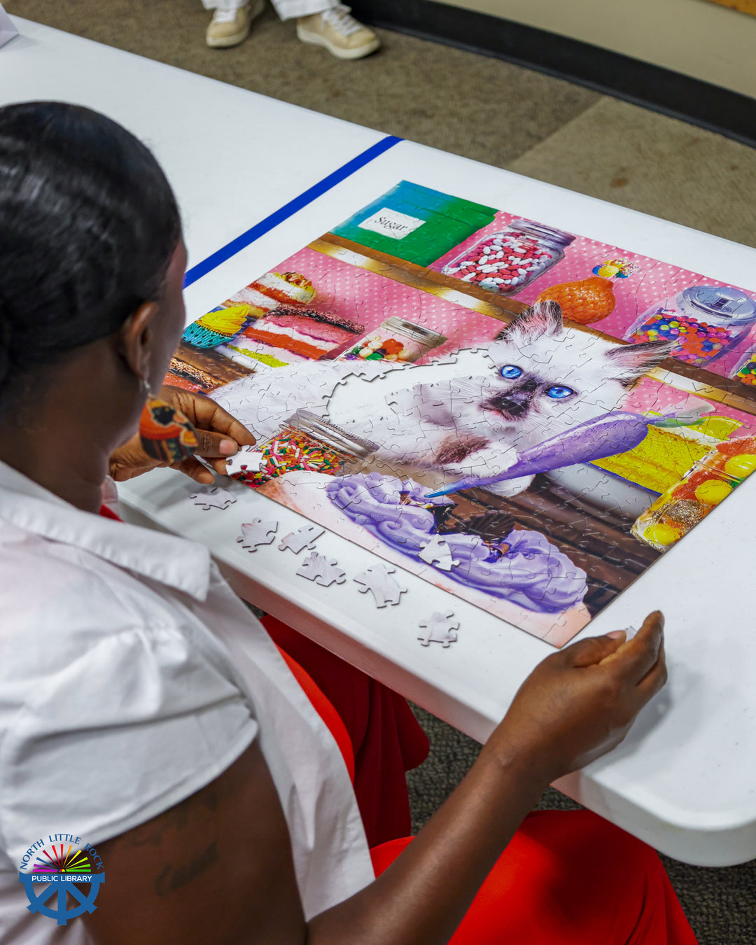 A seated woman looks down at an almost-finished puzzle of a cat frosting a cake.