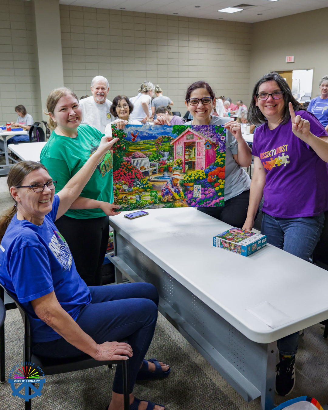 A group of 4 people hold up a completed puzzle with an image of a pink shed surrounded by colorful flowers.