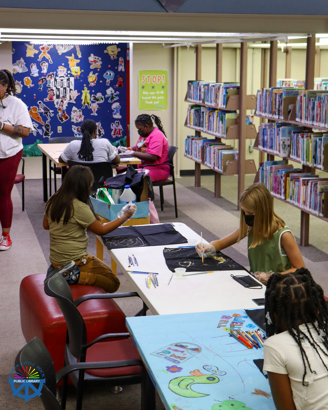 Tweens sitting at a table, wearing masks, painting with bleach on black shirts.