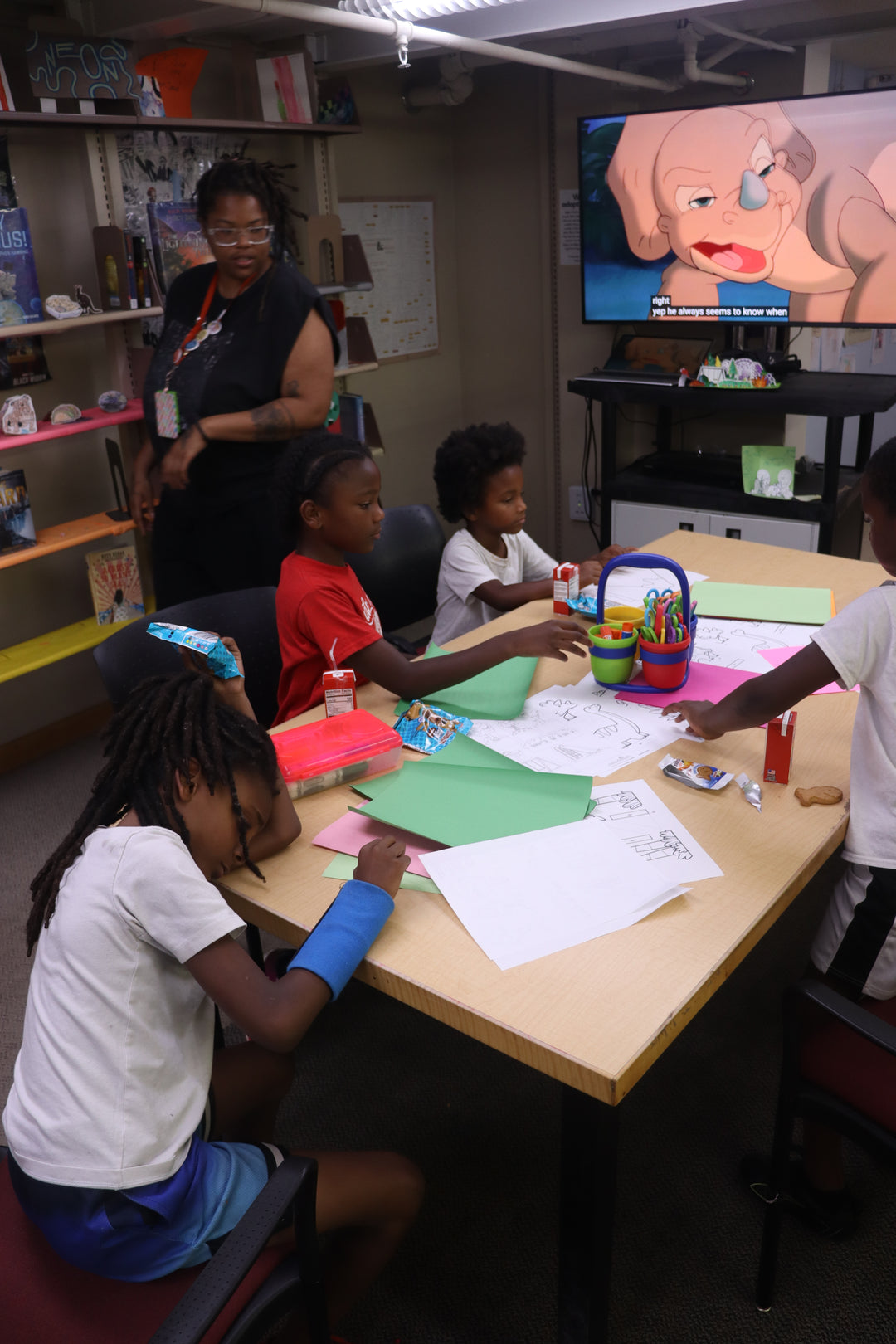 Library staff member Drienne watches a group of seated children make dinosaur dioramas while a movie plays on a television in front of them.