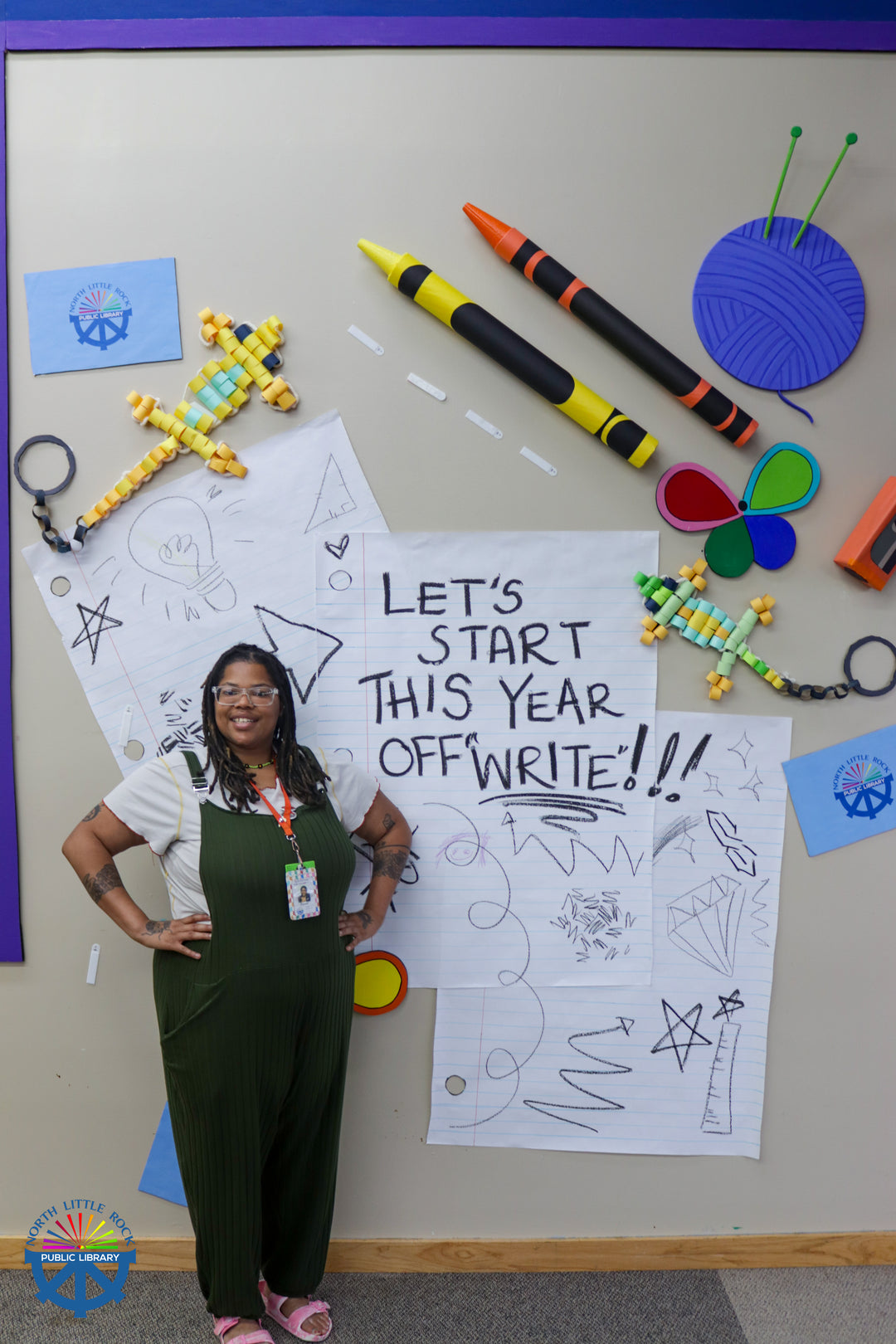 Tween specialist Drienne stands in front of a wall adorned with oversized school supplies, including a large piece of notebook paper reading Let's Start This Year Off "Write"!. 