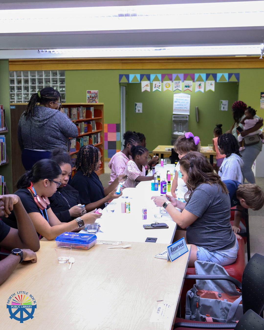 Tweens and families sit at a long table with paint and cups of water.