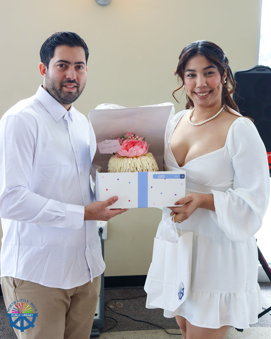 A couple dressed in white hold a decorated cake.