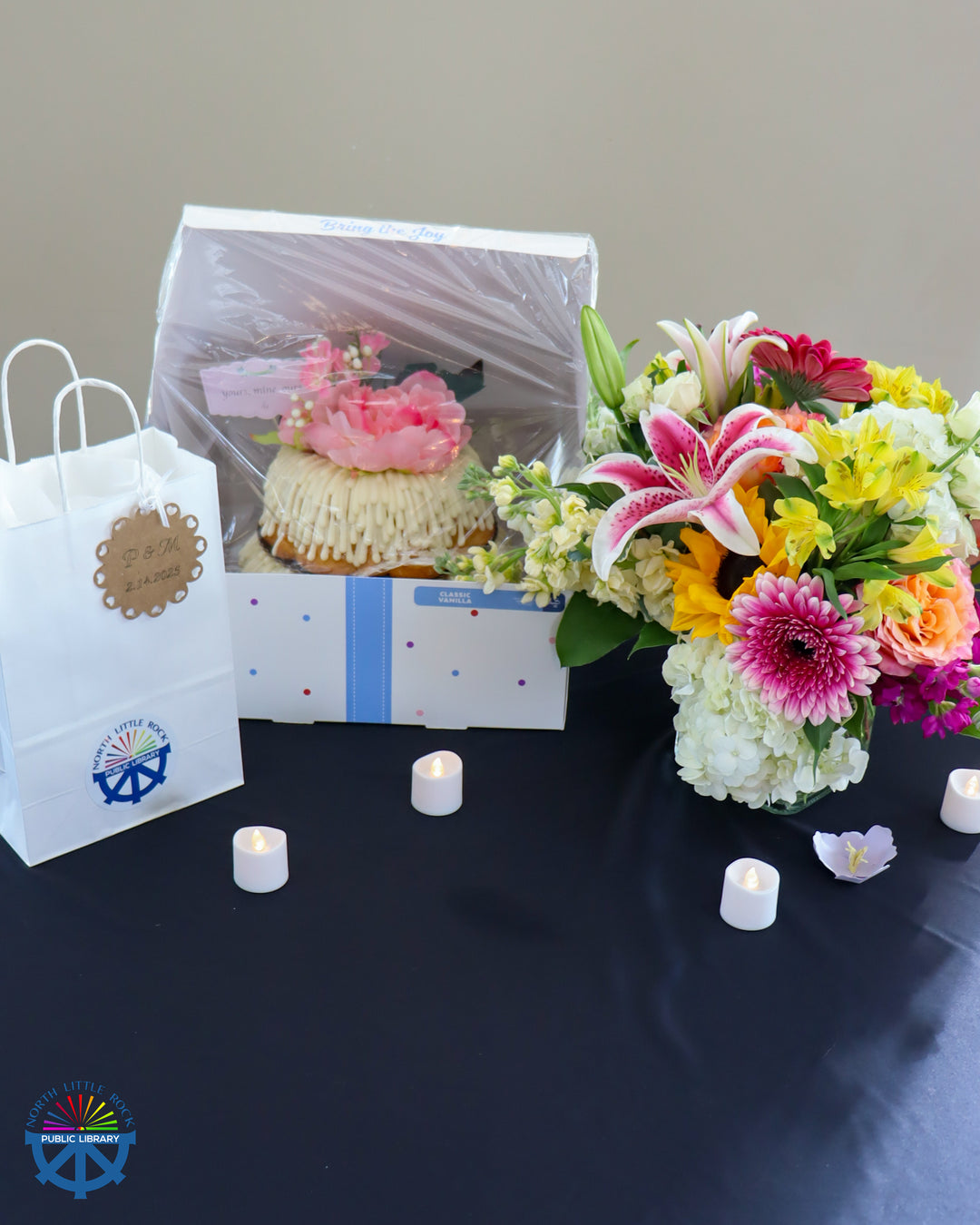 On a black table sits a white gift bag, a decorated cake, a floral arrangement, and electric candles.