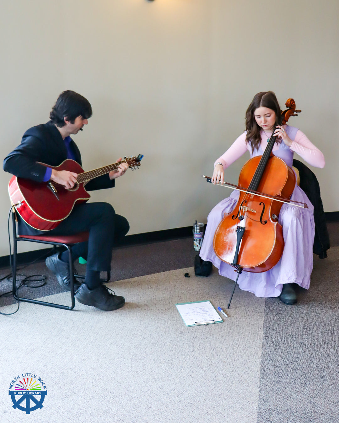 A man in a suit plays a guitar next to a woman in a lilac dress playing a cello.