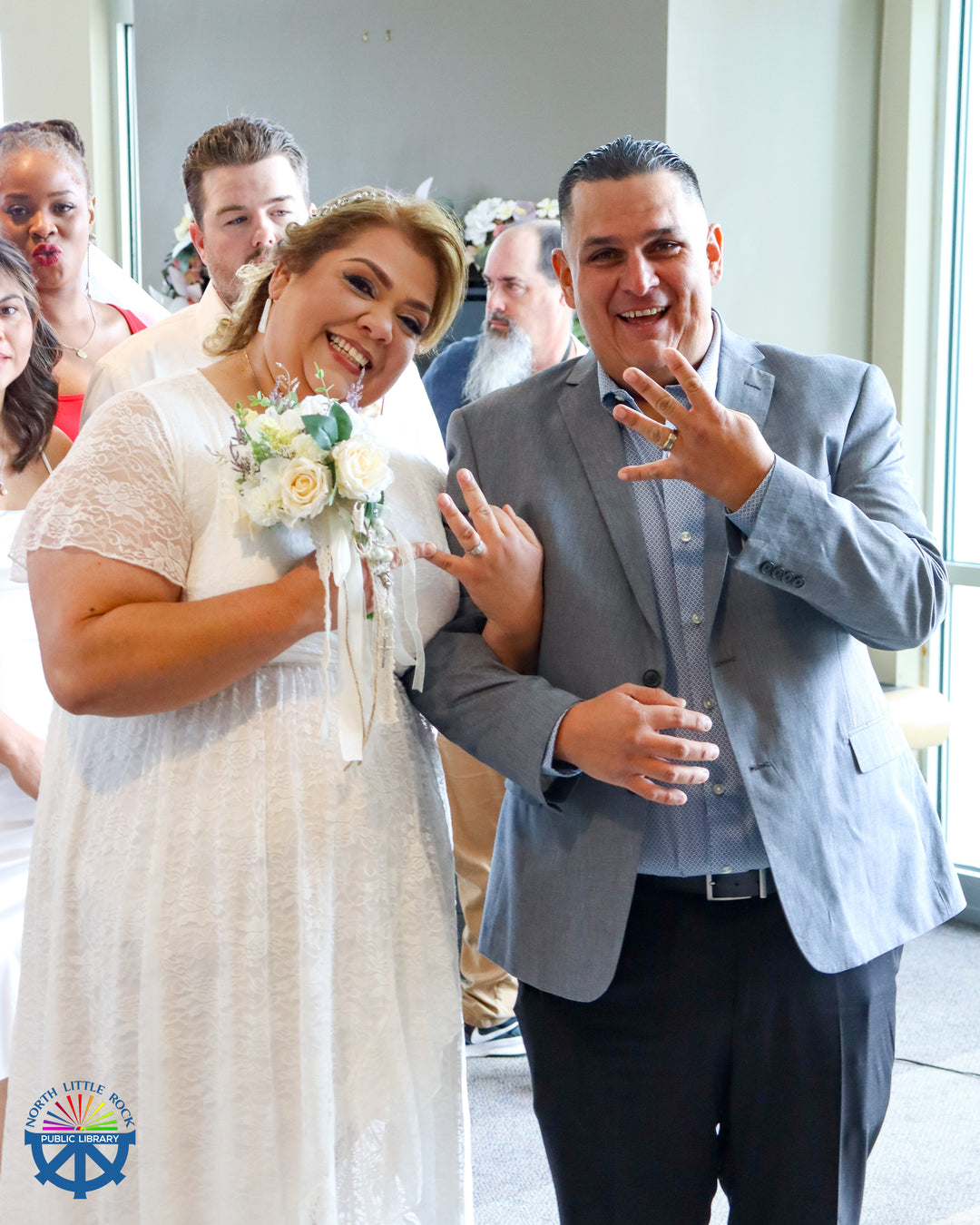 A man and woman wearing formal wedding clothes smile at the camera and show off their rings.