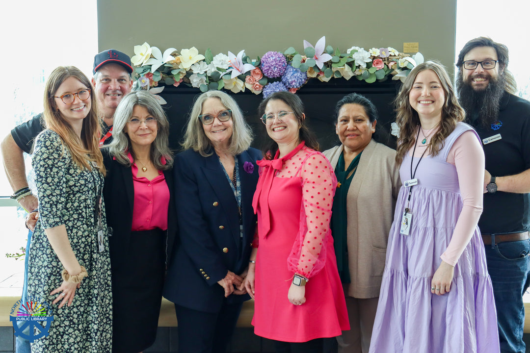 A group of 8 smiling library staff members pose in front of a mantle decorated with paper flowers.