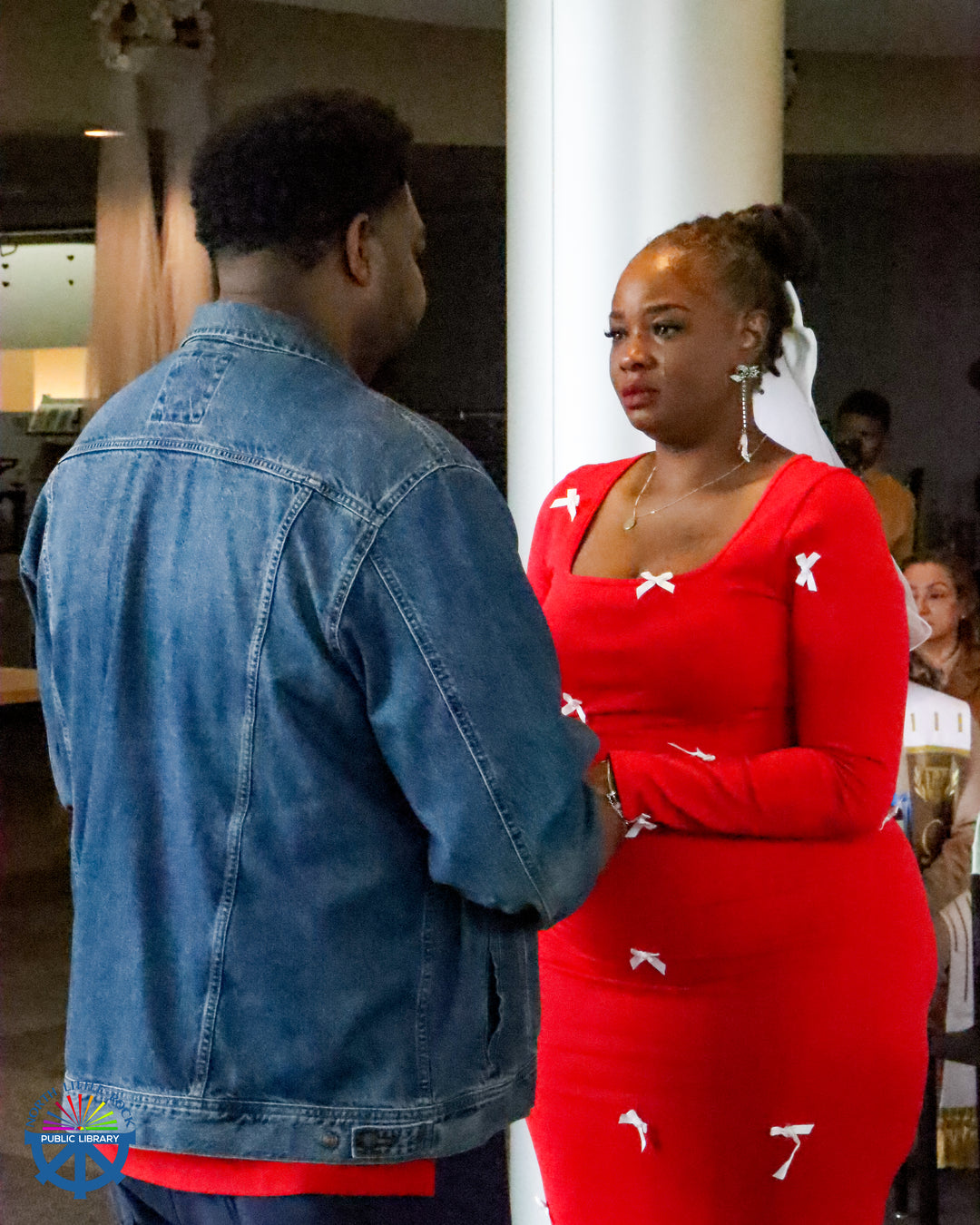 A woman wearing a red dress with white bows looks at a man wearing a denim jacket with tears in her eyes.
