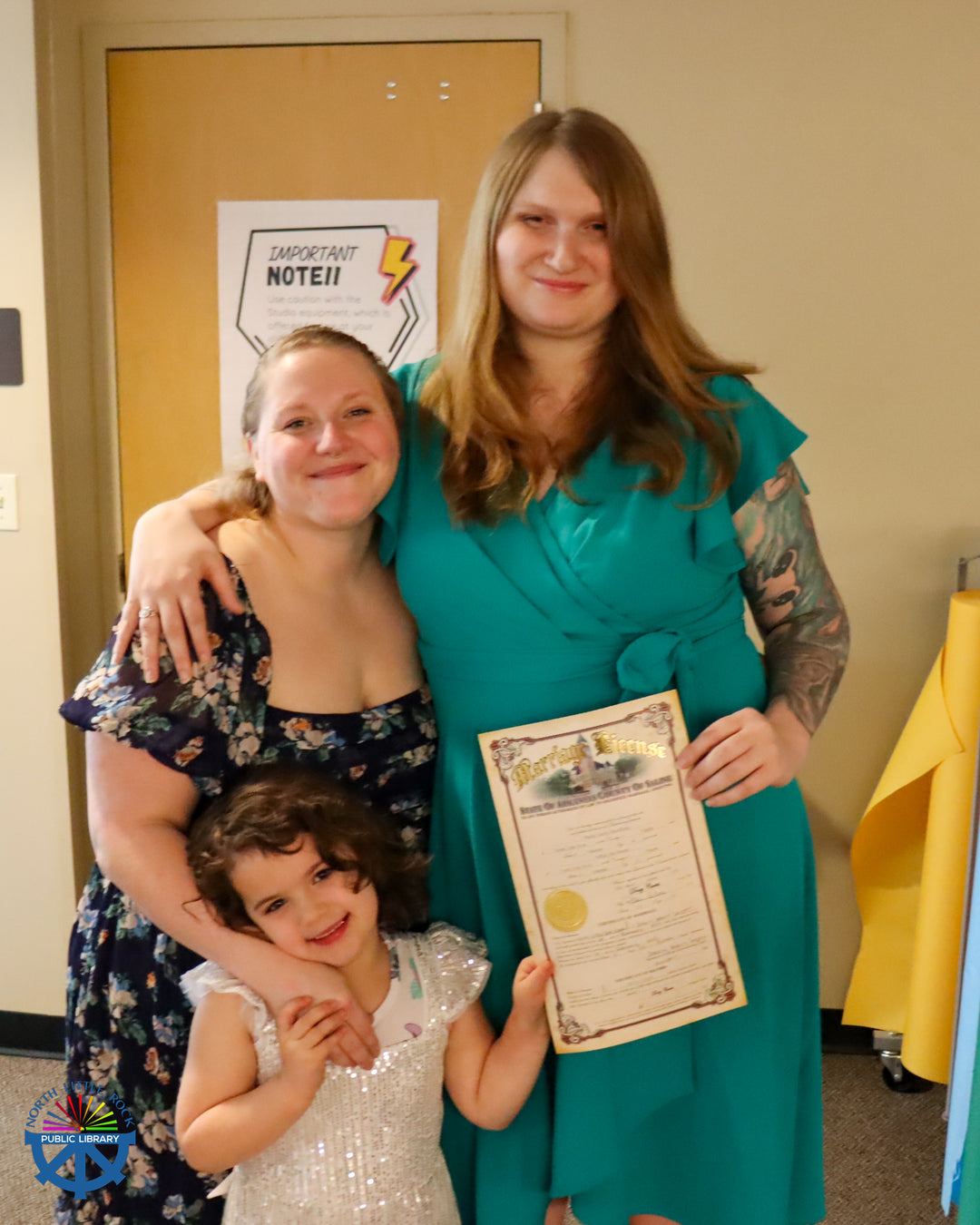 A smiling newly married couple in dresses pose with their marriage license and a young child.