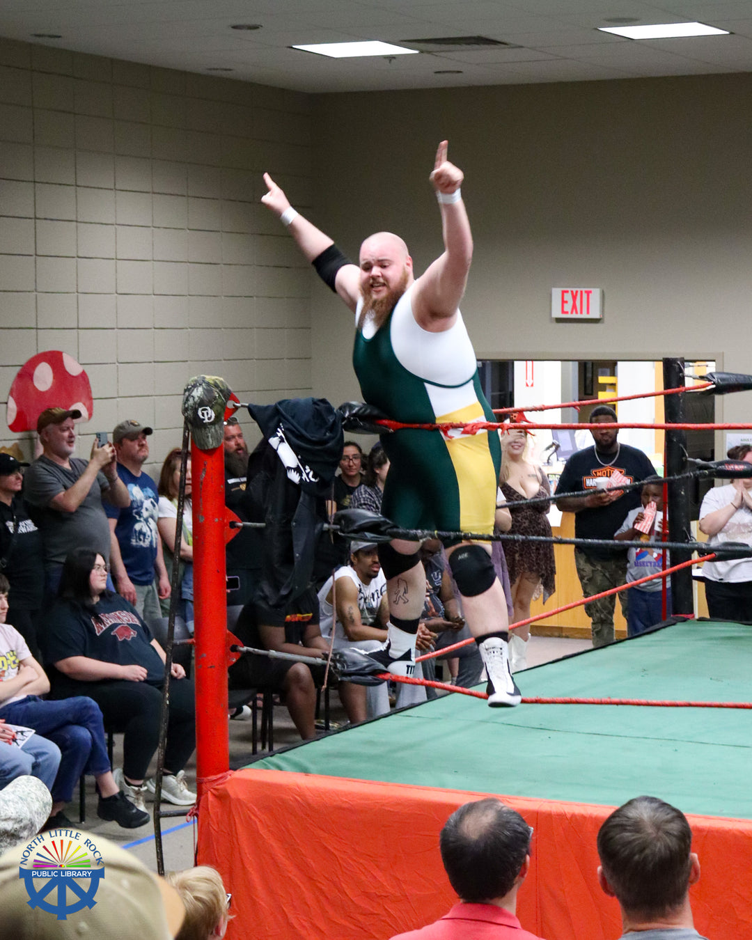 A wrestler in a white shirt and singlet stands on the ropes in the corner of the ring, arms raised towards the crowd below.