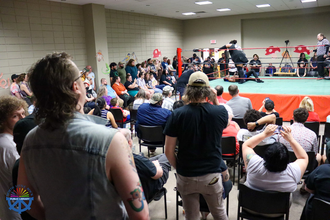 People sitting in chairs and standing around a wrestling ring as two wrestlers, one lying on his back and the other standing, engage in a match with a referee watching from across the ring.