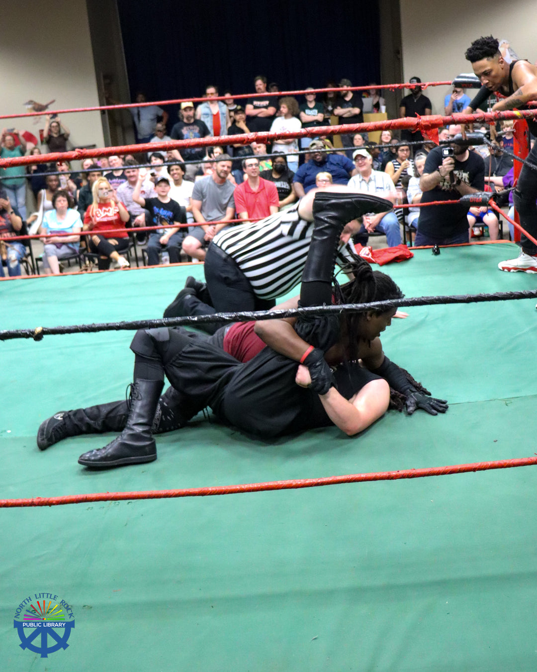 A referee slams his hand on the floor of the ring as one wrestler has another pinned. Around the ring are dozens of seated attendees.