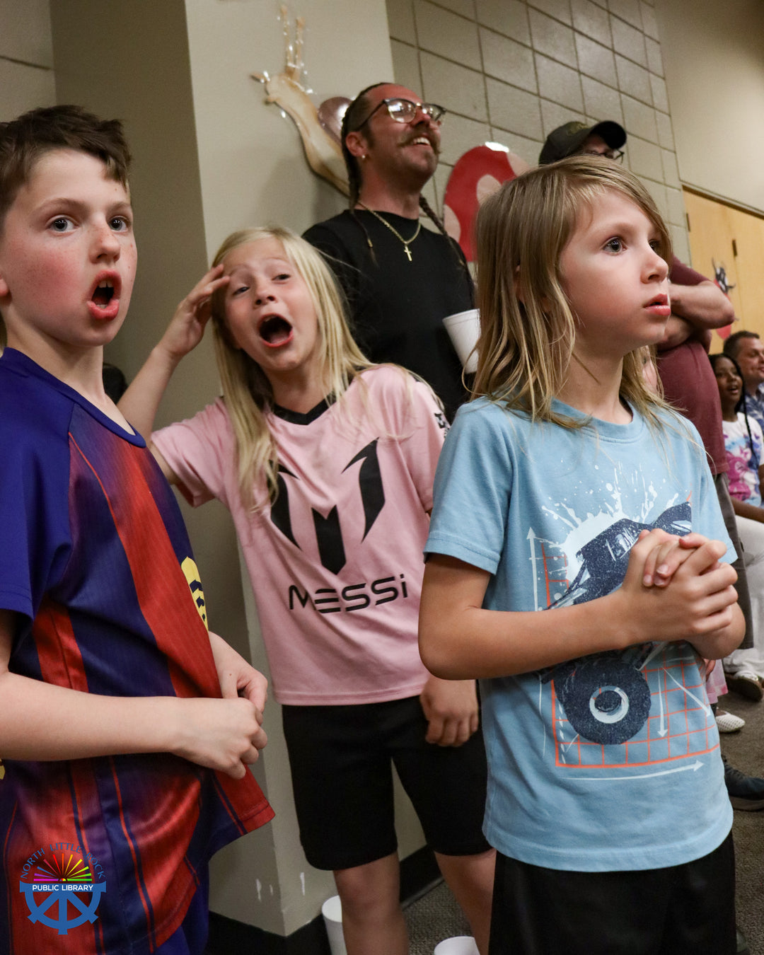 Three standing children watch the wrestling match with great interest and surprise. Adults stand and sit behind them, watching the action.