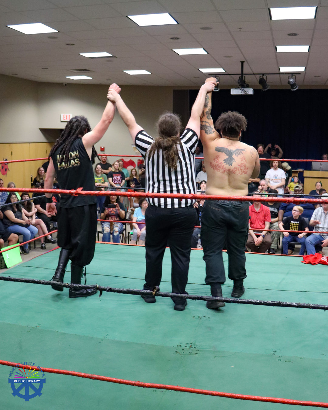 A referee in a striped shirt holds up the hands of two wrestlers, all three facing away from the camera and towards the attendees seated next to the ring.